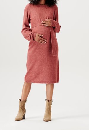 Pregnant woman wearing a rust-colored knit dress and tan ankle boots, standing with hands gently resting on her belly.