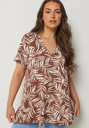 Vrouw die een bruin T-shirt met korte mouwen en een V-hals draagt, bedrukt met witte tropische bladeren, staand tegen een effen lichte achtergrond.