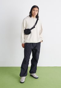 Haut à capuche beige clair, sac banane noir, pantalons noirs amples et baskets grises avec une semelle foncée, le tout sur un sol vert.