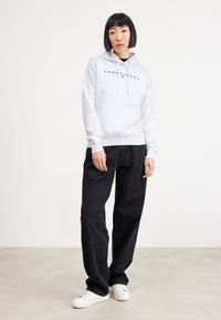 Felpa bianca con cappuccio e tasca frontale, con scritte nere. Abbinata a jeans neri larghi e scarpe da ginnastica bianche su uno sfondo neutro.