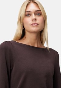 Jeune femme aux cheveux blonds raides portant un haut à manches longues marron foncé avec des fronces discrètes sur les épaules, sur un fond clair.