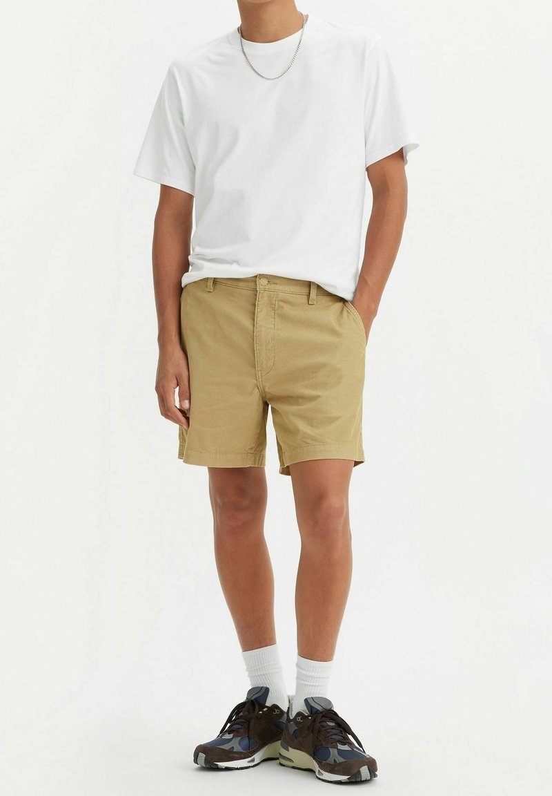 Homme portant un t-shirt blanc, un short beige, des chaussettes blanches, des baskets bleu foncé et marron, et un collier chaîne argenté, debout avec une main dans la poche.