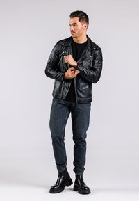 Giacca da motociclista in pelle nera con tasche con cerniera, indossata sopra una maglietta nera. Abbinata a jeans in denim scuro e stivali neri robusti.