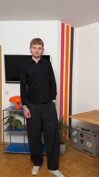 Άνδρας με μαύρο πουκάμισο και φαρδιά παντελόνια στέκεται σε σαλόνι με πορτοκαλί λάμπα, φυτά, τηλεόραση και ριγέ διακόσμηση στον τοίχο.