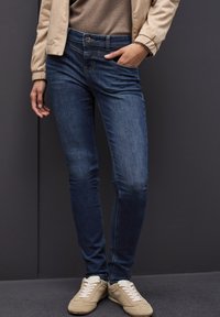 Tunna mörkblå jeans med en slät yta, fem fickor och knappstängning, parat med beige skor och en neutral jacka.