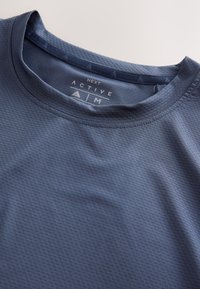 Gros plan d'un maillot de sport bleu texturé avec col rond et étiquette de taille imprimée "Next Active M" à l'intérieur du col.