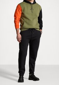 Mężczyzna w kolorowym hoodie z pomarańczowymi i czarnymi rękawami, oliwkowym korpusem, czarnych joggerach i czarnych sneakersach, stoi z rękami w kieszeniach.