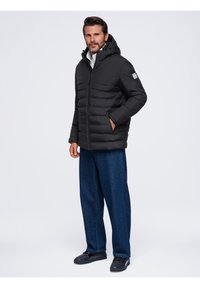Homme portant une doudoune noire avec capuche, un jean bleu foncé et des chaussures noires, debout avec une main dans la poche sur fond blanc.