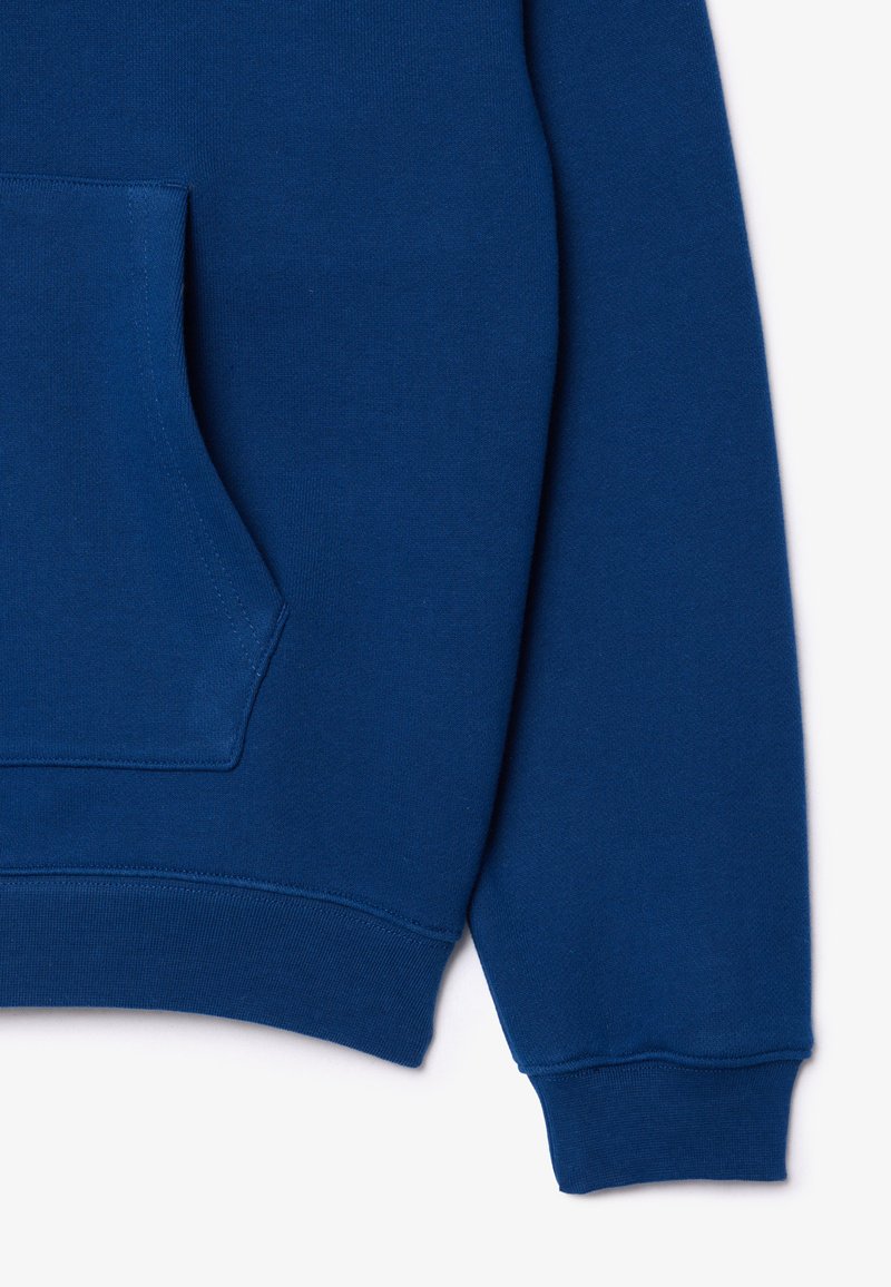 Sudadera de algodón azul con bolsillo frontal, puños y dobladillo acanalados. La textura es suave con un acabado liso. El diseño es simple y casual.