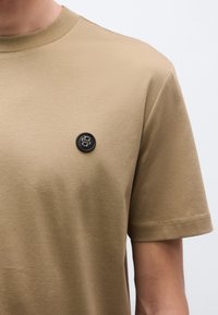 Puha anyagból készült khaki színű póló, kör alakú nyakkivágással és rövid ujjakkal, a mellkason fekete kör alakú logóval.