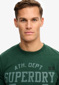 T-shirt en coton vert avec un col rond et un texte imprimé "SUPERDRY" en gris clair avec des accents noirs sur l'épaule droite.