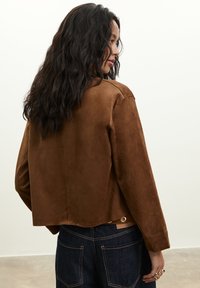 Žena s dlhými vlnitými čiernymi vlasmi, oblečená v hnedej semišovej bunde a tmavých denimových džínsoch, otočená bokom na obyčajnom pozadí.