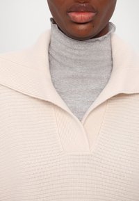 Lys beige strikkegenser med bred, brettet krage over en grå ribbestrikket høyhalser. Teksturert stoff med vertikalt mønster og V-utsnitt.