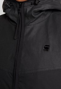 Black hooded jacket made of textured fabric. Features a front zipper, adjustable drawstrings, and a small logo on the chest.