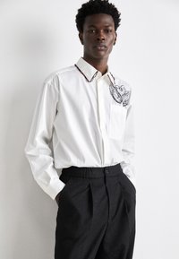 White long-sleeve shirt with red and black embroidered collar and a chest pocket, paired with black textured trousers featuring a logo pattern.
