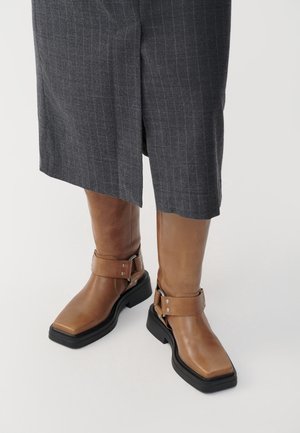 Bottines en cuir marron avec semelles épaisses noires, brides à boucle argentée, portées avec une jupe à rayures grises à fente frontale.
