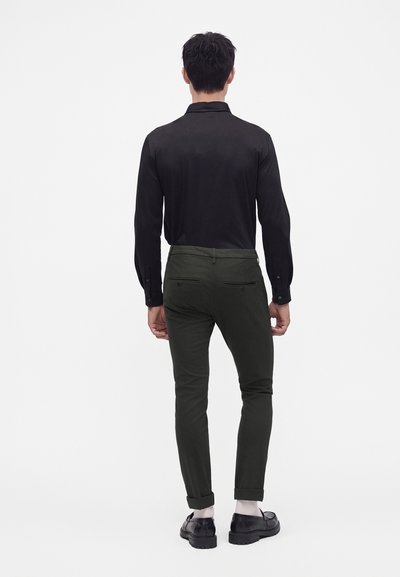 Homme portant une chemise noire à manches longues, un pantalon slim vert foncé et des mocassins noirs, debout de dos sur un fond uni.