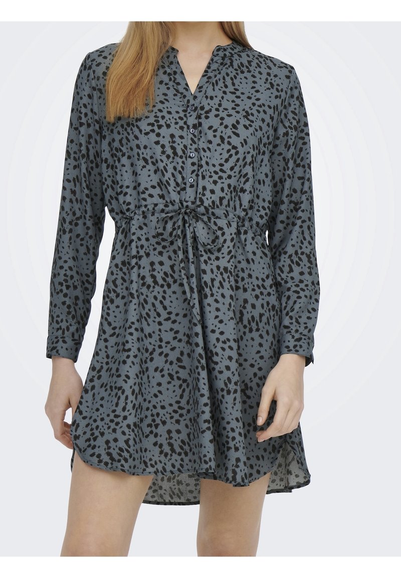 Femme portant une robe grise avec des taches noires irrégulières, des manches longues, un col boutonné et une ceinture nouée à la taille.