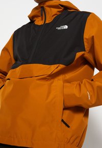 Homme portant une veste The North Face orange et noire avec zip avant et poches pour les mains, l'étiquette sur la manche montre la technologie du tissu DryVent.