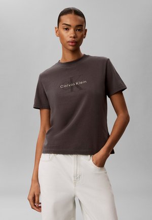 Jonge vrouw die een donkergrijs Calvin Klein T-shirt en witte broek draagt, met één hand in de zak, tegen een effen grijze achtergrond.