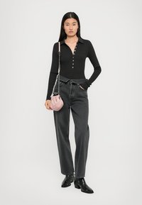 Woman wearing black long-sleeve buttoned top, high-waisted dark gray jeans, black ankle boots with studs, holding a pink handbag.