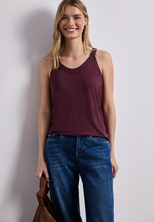 Uśmiechnięta kobieta w bordowej bluzce bez rękawów i niebieskich dżinsach, trzymająca brązową torebkę na tle jednolitej jasnej ściany.