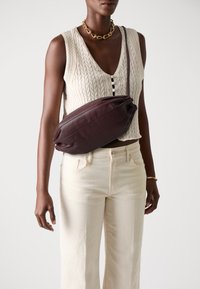 Borsa in pelle bordeaux scuro con una forma ovale elegante, indossata a tracolla. Il top in maglia testurizzata color crema presenta un profondo scollo a V e bottoni decorativi.