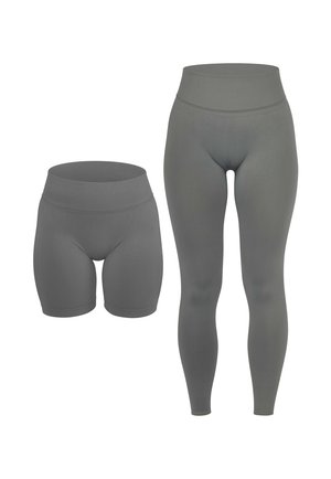 Frau trägt graue Leggings mit hohem Bund und weiße Turnschuhe, steht mit entspannten Händen seitlich vor einem schlichten Hintergrund.