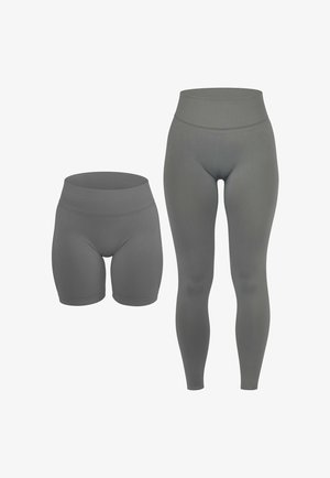 Frau trägt graue Leggings mit hohem Bund und weiße Turnschuhe, steht mit entspannten Händen seitlich vor einem schlichten Hintergrund.