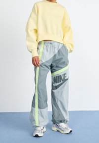 Person som bär en gul kortärmad sweatshirt, grå Nike vindjacka med neonränder, och vita sneakers som står på ett blått golv.