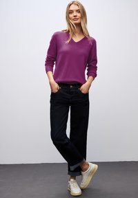 Pull violet en V, coupe décontractée. Jeans bleu foncé avec les poignets retroussés. Baskets blanches avec semelles beige. Arrière-plan minimaliste.
