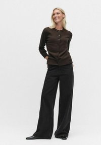 Brun stickad cardigan med rund halsringning och knappstängning, tillsammans med svarta vida byxor som har en subtil textur och lyster.