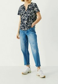 Blusa a maniche corte a motivi blu e bianchi con scollatura a V, abbinata a jeans di denim blu e sneakers grigie. Taglio informale con orlo arrotondato.