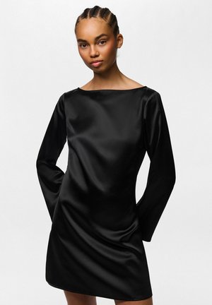 Jeune femme aux cheveux tressés portant une mini-robe noire en satin à manches longues, debout devant un fond clair uni.