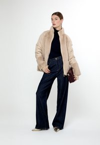 Giacca in pelliccia sintetica beige con colletto alto, abbinata a jeans a gamba larga blu scuro. Il modello tiene una clutch bordeaux; tacchi a punta dorati visibili.