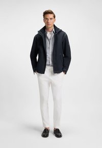 Tamsiai mėlynas švarkas su gobtuvu, priekyje užtrauktuku, matomos siūlės, derinamas su šviesiai pilka marškinėlių palaidine su sagutėmis ir baltomis kelnės. Juodi batai užbaigia įvaizdį.