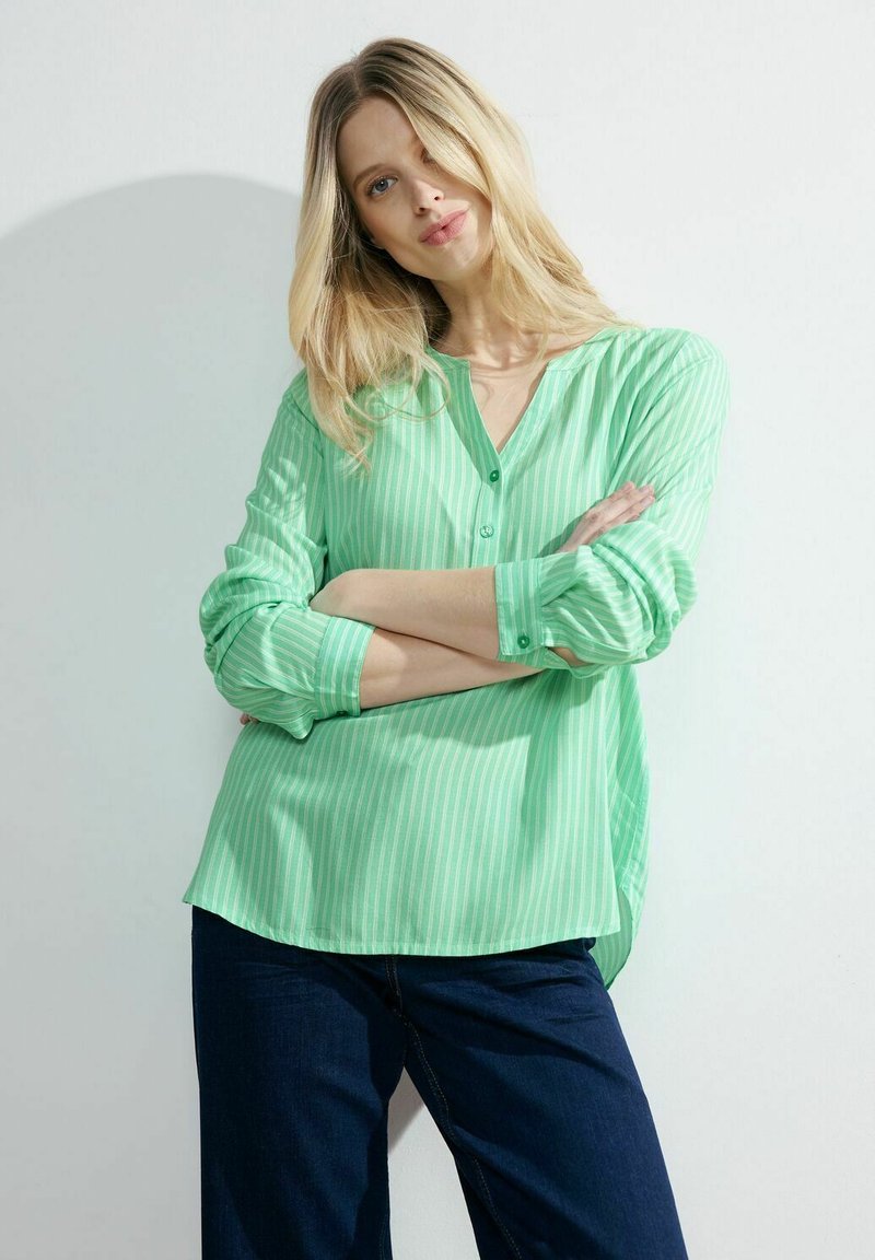 Blusa verde a righe con scollatura a V e bottoni, maniche lunghe arrotolate, abbinata a jeans a gamba larga blu scuro. Tessuto dalla consistenza liscia.