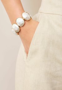 Guldtonet armbånd med tre ovale, facetterede hvide sten, båret på et håndled, delvist synligt i en beige lomme af et let stof.