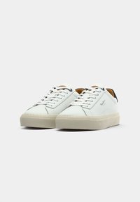 Baskets en cuir blanc avec talon noir contrastant, bout rond et semelle en caoutchouc beige. Elles possèdent des lacets blancs et un logo discret sur le côté.