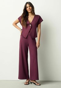 Bordový voľný overal s krátkymi rukávmi a výstrihom do V, zdobený dekoratívnym gombíkom. Hladká, mäkká textúra a široké nohavice.