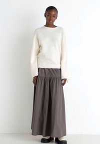 Bílý oversized svetr s dlouhými rukávy, kombinovaný s hnědou maxi sukní s nabíraným pasem, obutý s černými platformovými botami.