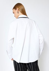 Camisa blanca de manga larga con un collar negro en forma de ondas, corte holgado y textura suave; el modelo lleva pantalones rayados negros.