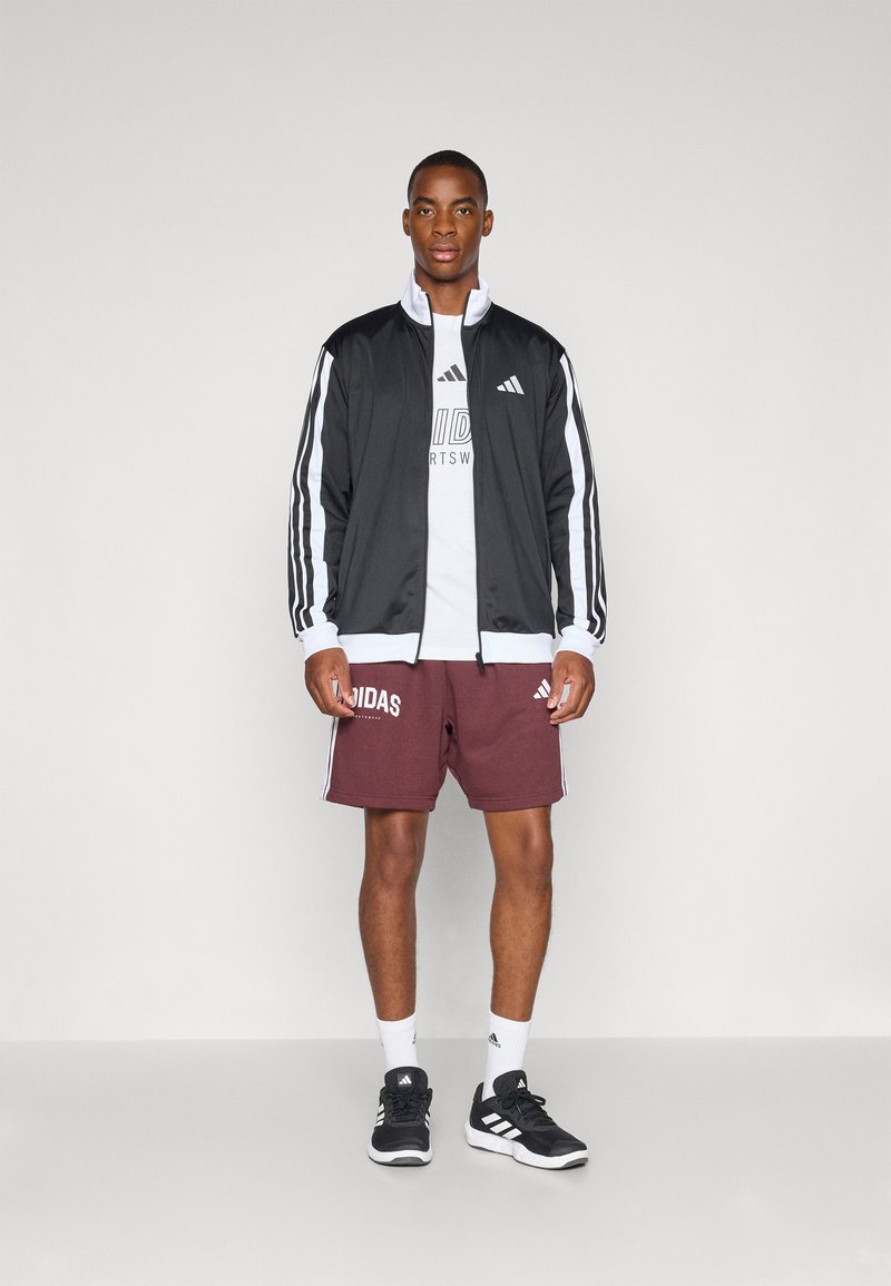 Hombre atlético vistiendo una chaqueta de chándal Adidas en negro y blanco, camiseta blanca con logo, pantalones cortos marones de Adidas, calcetines blancos y zapatillas negras de Adidas.