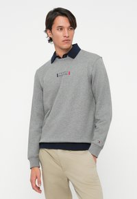 Noor mees kannab halli Tommy Hilfiger spordipusa, mille all on ninaga särgi, beežid püksid, seistes ühe käega taskus valge tausta taustal.