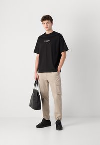 Zwarte katoenen t-shirt met logo, beige cargobroek met zijzakken, zwarte sneakers en een zwarte totebag. Effen achtergrond.