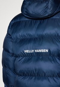 Doudoune bleu marine en nylon, avec un col montant et un logo blanc "HELly HANSEN". Comprend des sections matelassées pour l'isolation.