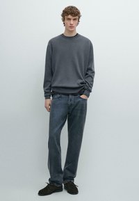 Gray sweater with ribbed cuffs and hem, paired with dark denim jeans. Model stands with hands in pockets, wearing dark shoes. Simple layout.