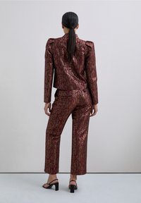 Mujer con coleta negra larga vestida con un blazer brillante de patrones en marrón y dorado y pantalones a juego, de pie de espaldas sobre un fondo claro.