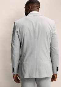 Chaqueta de traje gris claro hecha de tejido texturizado, con solapas de muesca, puños abotonados y un corte entallado. La vista trasera muestra líneas limpias.