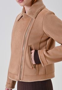Giacca in morbido suede taupe con colletto in pelliccia, zip laterale e dettagli a costine sui polsini. Presenta una tasca laterale e un design pulito e strutturato.